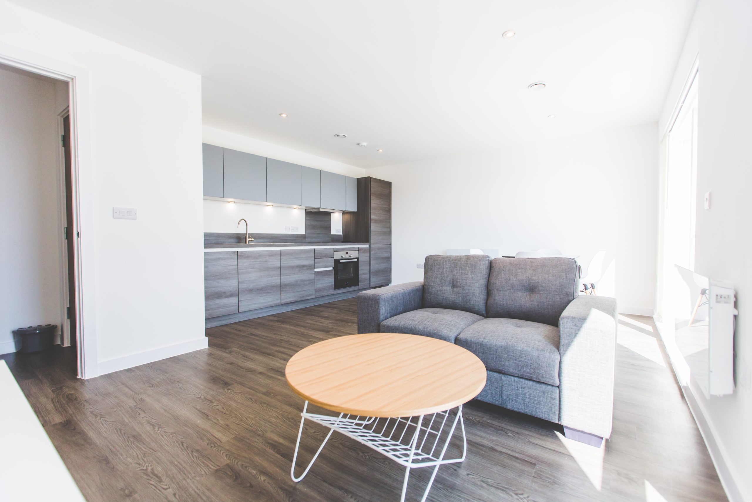 Lose up view of a grey double sofa, light circular coffee table with a grey kitchen in the background.