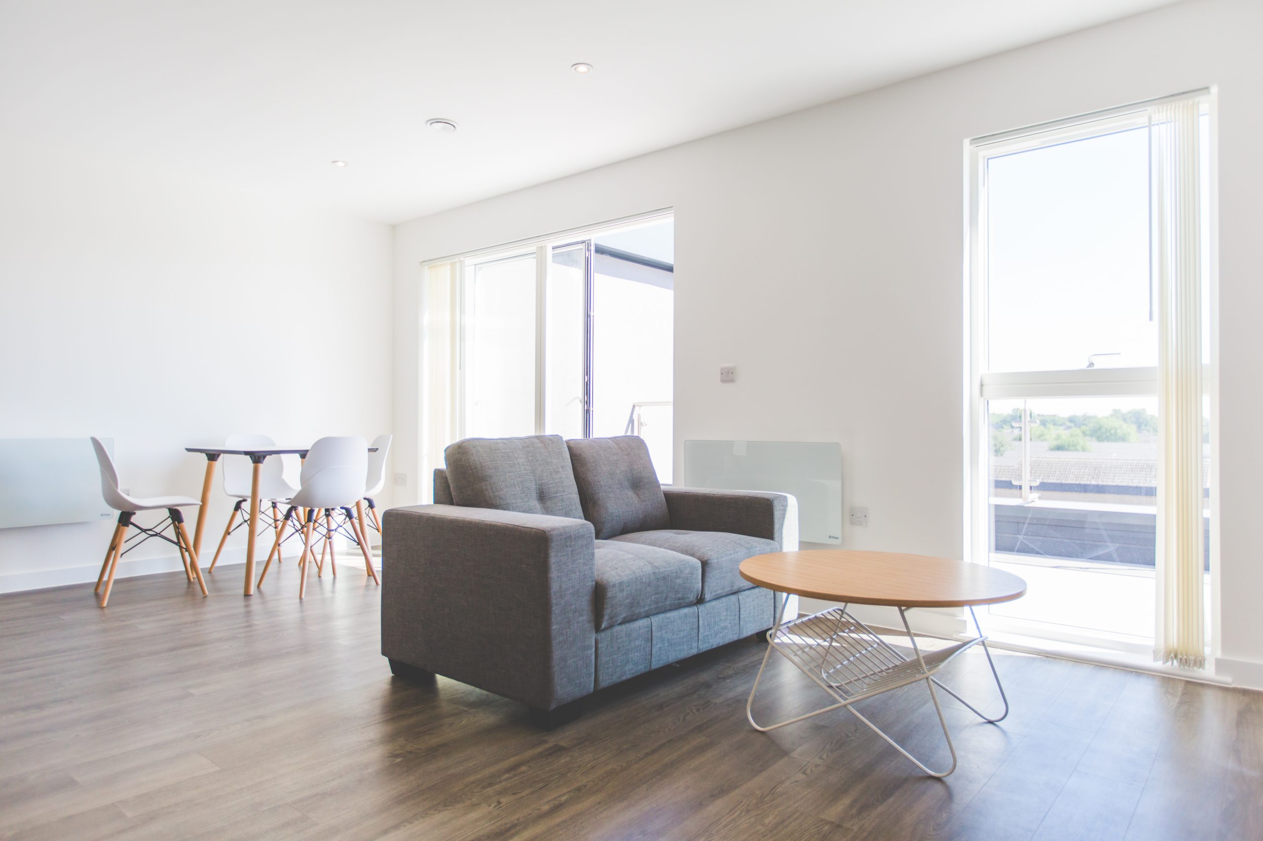 Interior view of a large open plan lounge with dark wooden floor, two large windows a grey double sofa and white table and chairs in the background.