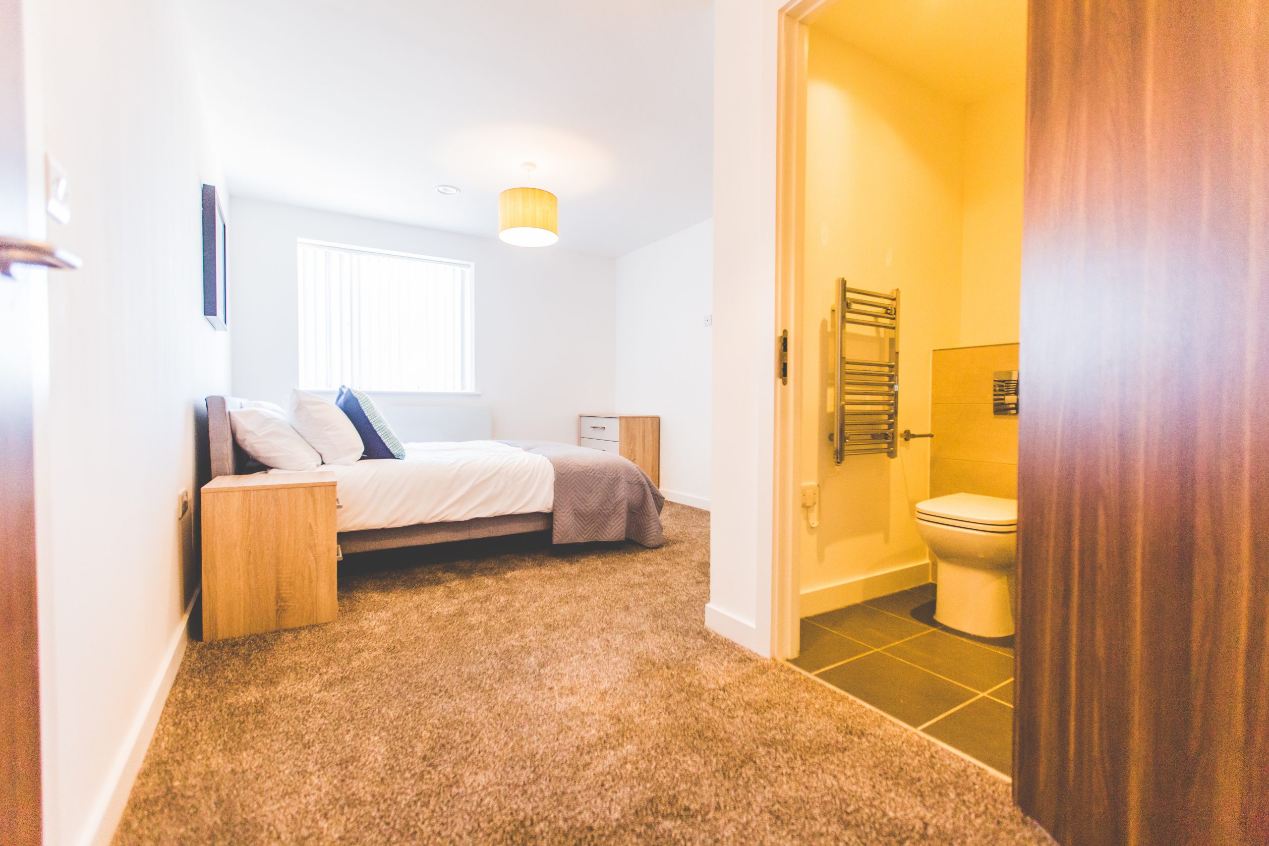 Interior view of a bedroom with light brown carper, large white bed and an en-suite to the right.
