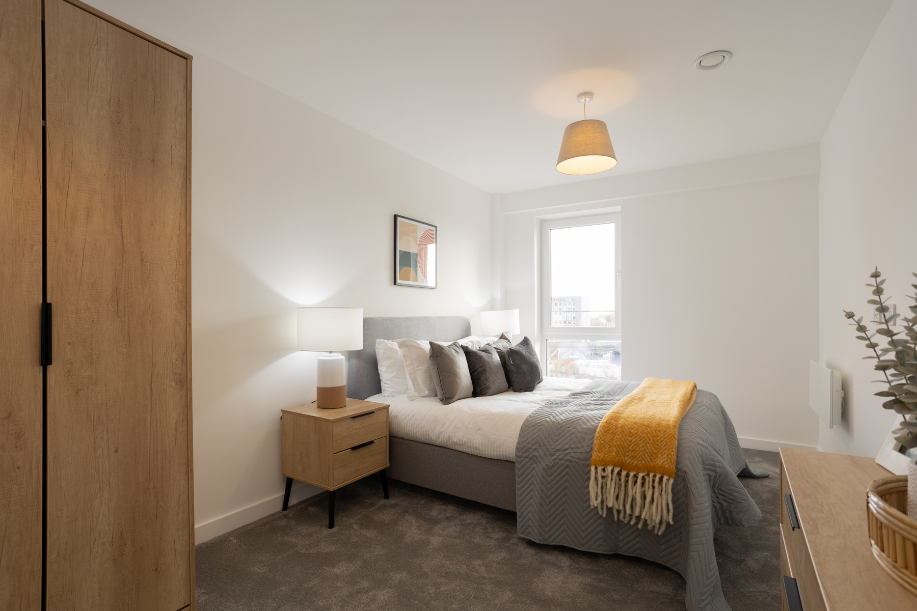 Bedroom with grey carpet, grey bed, white sheets and wooden wardrobe to the left.