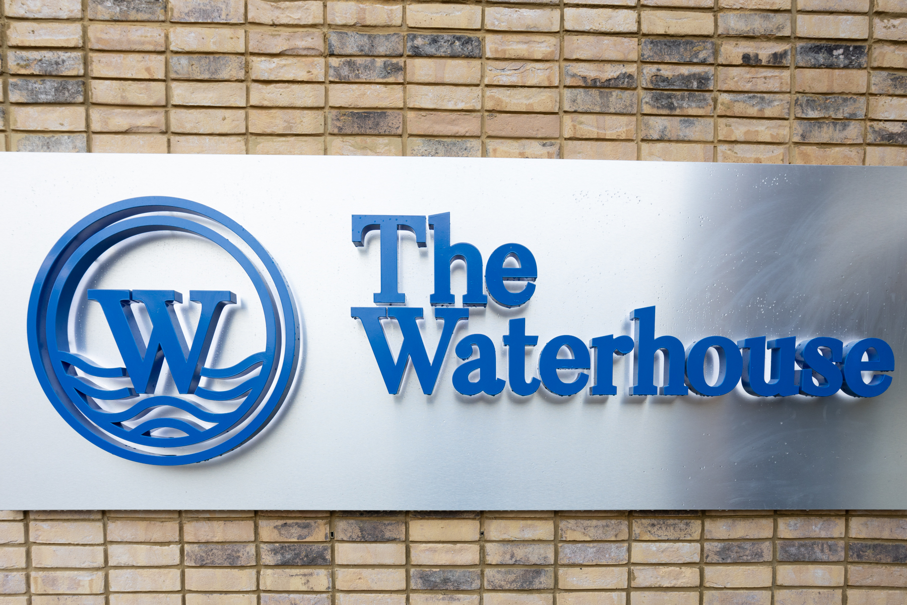 Exterior sign of The Waterhouse, brushed chromed metal background with the blue W logo and name.