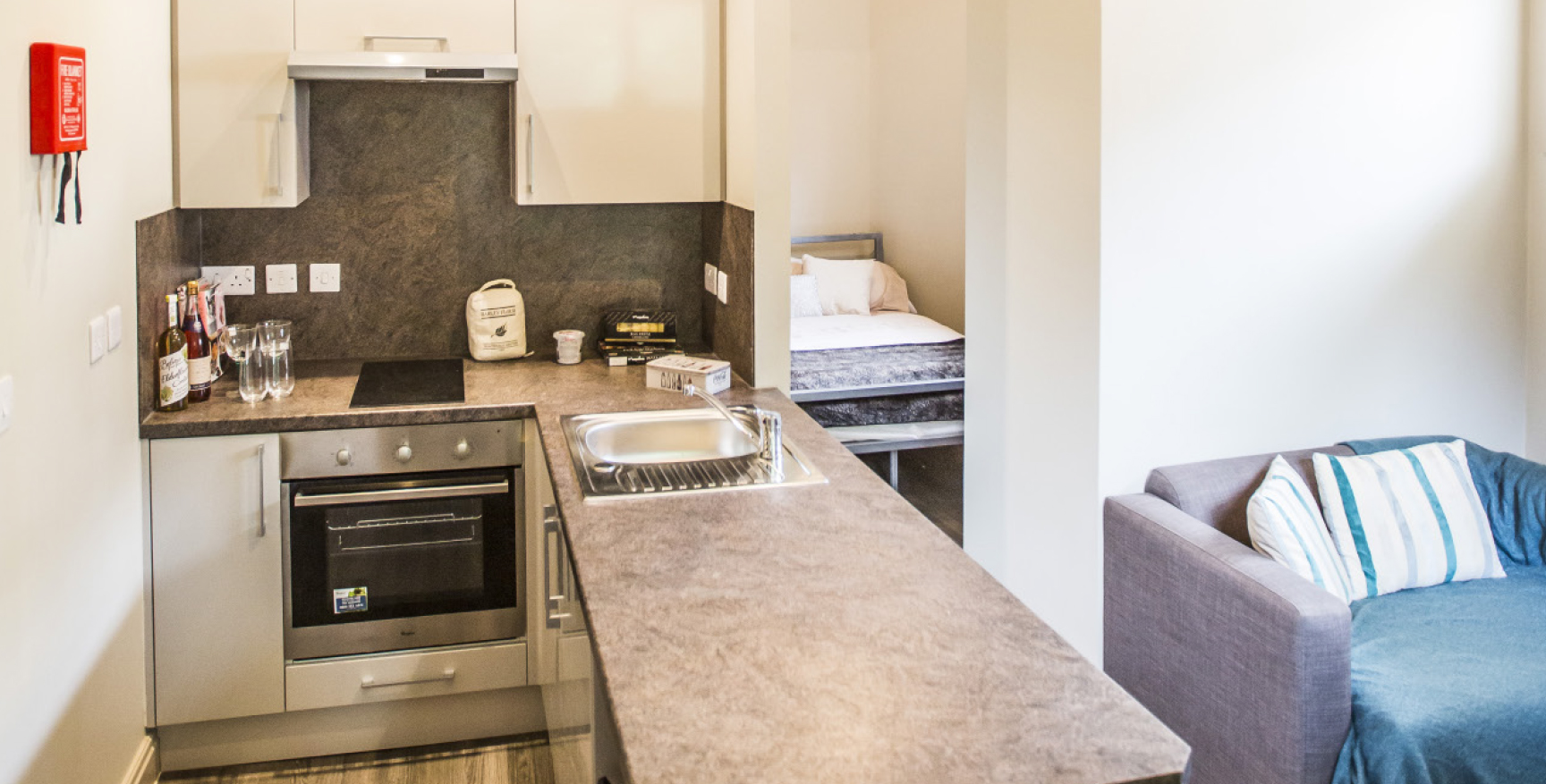 Kitchen with and grey peninsula worktop, single green chair to the right and a bedroom in the background.