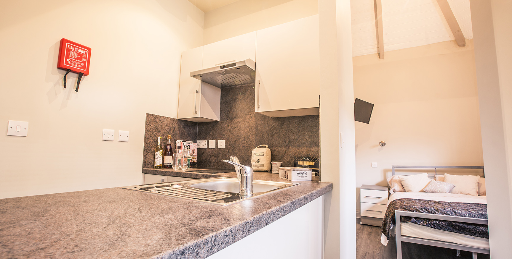 Close up view of the kitchen with a fire safety blanket attached to the wall and a double bed in the background.