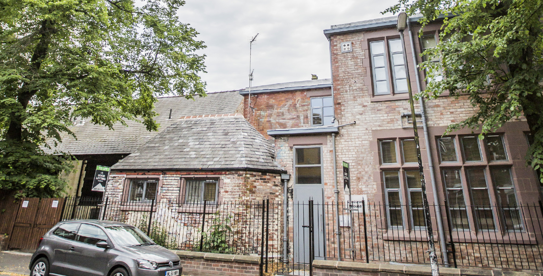 Exterior view of Landcoss House with a small grey car parked at the front, large green trees either side of the picture and a brick building with an iron fence at the front.