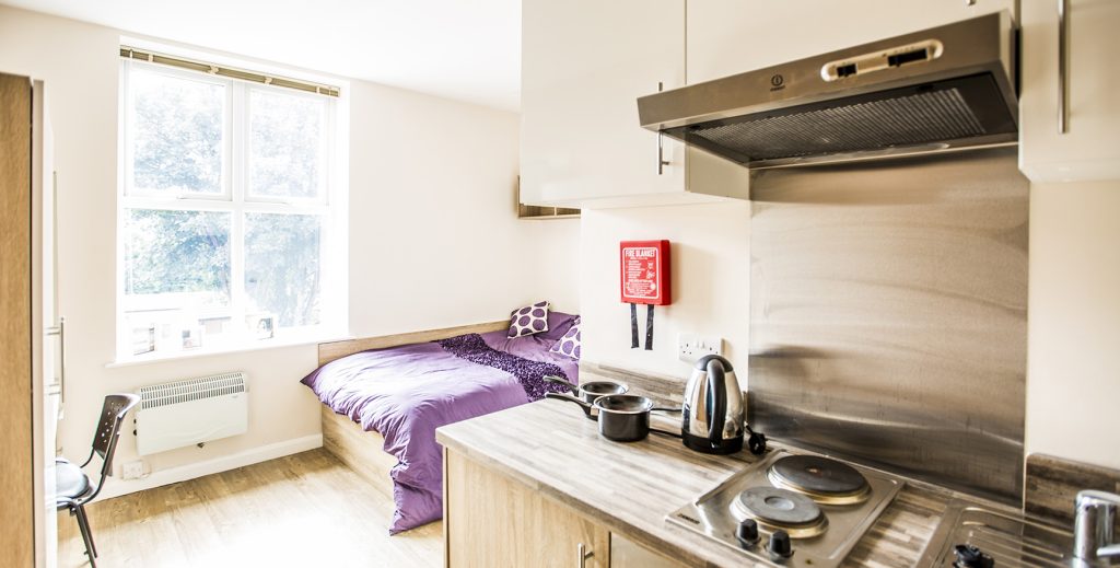 View of student room showing small kitchen unit with double bed in front of a large window.