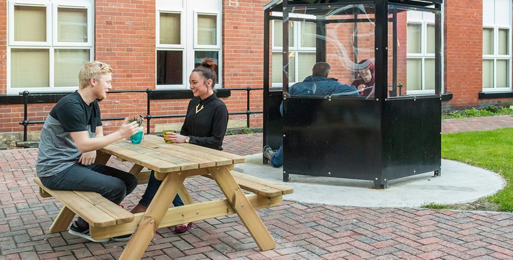 Exterior communal view of a picnic bench with a male and female student sat down. Along side is a covered small area with two students talking.