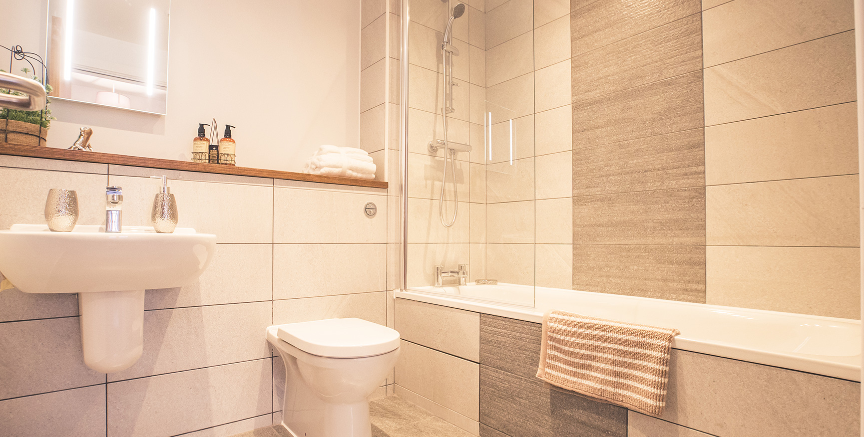 Level view of a bathroom with sink, toilet and bath with white tiles and an illuminated mirror.