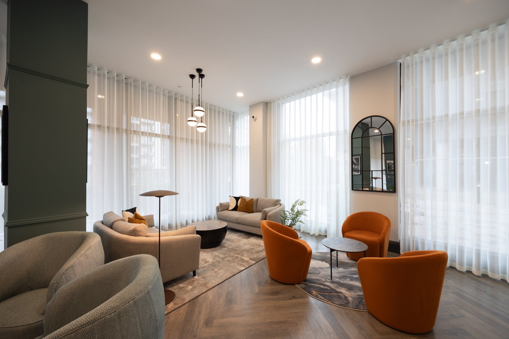 Communal area with floor to ceiling sheer curtains, wooden floor, three orange arm chairs and cream sofas.