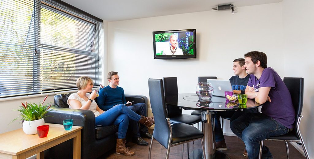Male amd female sat on leather sofa in front of a TV whilst chatting to two male students sat at a dining table.
