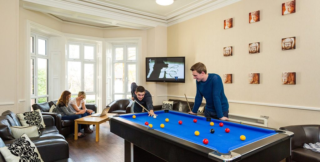 Two male student playing pool whilst two female students chat on a sofa.
