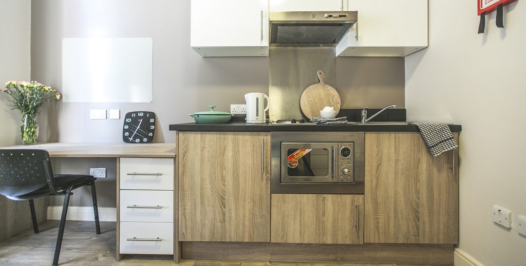 View of desk and chair along side a kitchen with white wall units and wooden veneered doors below.