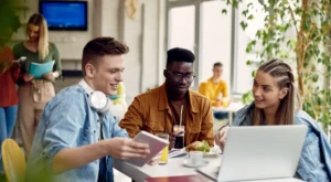 Three student sat at a table in a student cafe chatting and looking at a laptop. Student Accommodation Leeds Bolton York